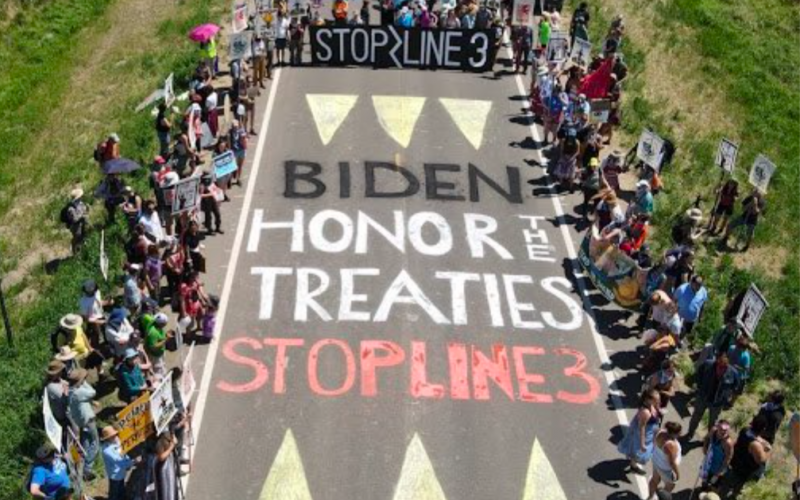At the Treaty People Gathering, hundreds gather around road art that says "Biden - Honor The Treaties, Stop Line 3"