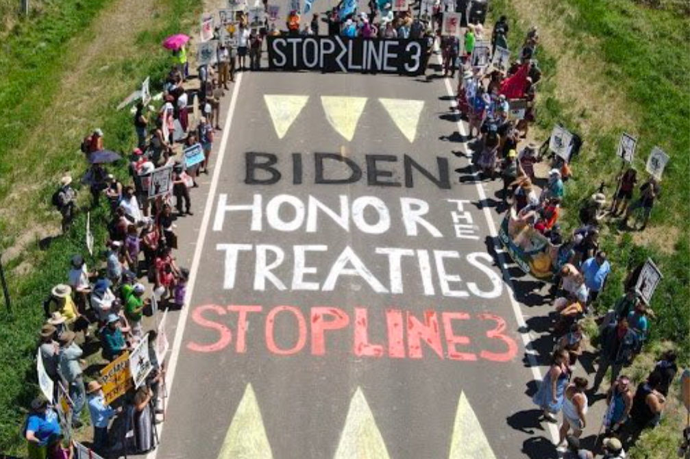 At the Treaty People Gathering, hundreds gather around road art that says "Biden - Honor The Treaties, Stop Line 3"