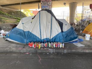 Candles are lined up in front of a blue and white tent under a freeway overpass in Oakland.