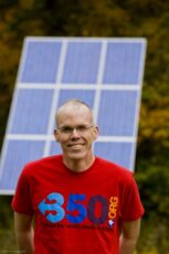 Photo of Bill McKibben standing in front of a solar panel, wearing a read 350.org shirt.
