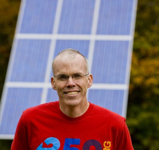 Photo of Bill McKibben standing in front of a solar panel, wearing a read 350.org shirt.