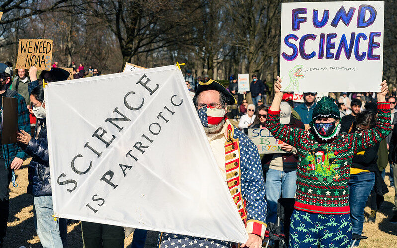 Protestors in colorful gear hold up "Science is Patriotic" and "Fund Science" posters at the Stand up For Science protest in Washington DC in March 2025.