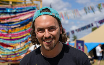 Sam Furness is a white male with brown hair pulled back under a backwards turquoise baseball cap. He is wearing a black T-shirt with a colorful logo and smiling warmly directly at the camera. In the background are tens and colorful banners of a festival.