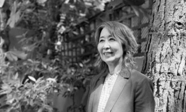 Black and white photograph of Haru Yamada, a Japanese woman with shoulder length hair. Haru is wearing a white shirt with blazer and is smiling at something to the left of the camera. She is standing in front of a large tree with dense foliage behind her.
