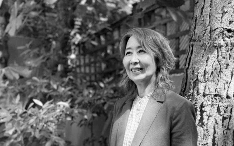 Black and white photograph of Haru Yamada, a Japanese woman with shoulder length hair. Haru is wearing a white shirt with blazer and is smiling at something to the left of the camera. She is standing in front of a large tree with dense foliage behind her.