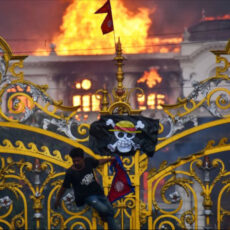 a photo of someone climbing the gilded gates of the Nepalese parliament building, hanging a flag with a skull and crossbones and wearing a straw hat, with the parliament burning in the background