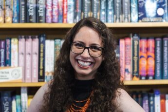 Ashley Kirsner is white woman with brown curly hair and glasses. She smiles at the camera in front of a book shelf.