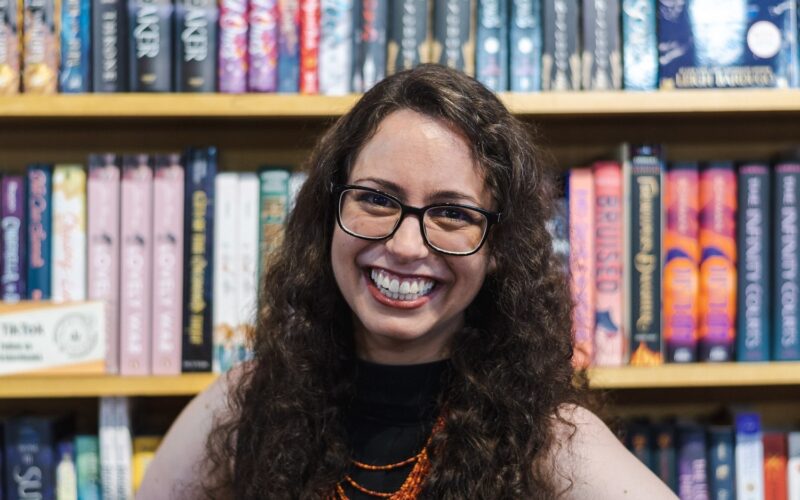 Ashley Kirsner is white woman with brown curly hair and glasses. She smiles at the camera in front of a book shelf.
