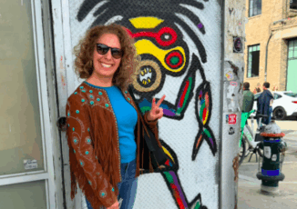 Estee Levine is a white woman with curly hair. She stands in front of a colorful mural by DC artist Reggie "Art" Gillumo