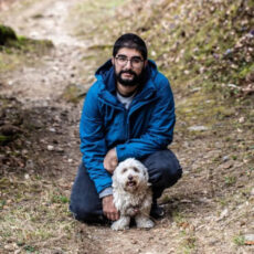 a photo of Elia Ayoub crouching in a blue jacket in front of a wooded trail with his dog