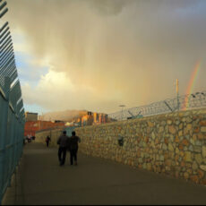 a photo from the El Paso / Juarez border showing walles, fences, barbed wire, border agents, a passing rainshower and rainbow