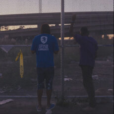 A still from the film showing two figures from behind, leaning up against and peering through a fence at an empty lo next to highway interchanges under a purplish sky