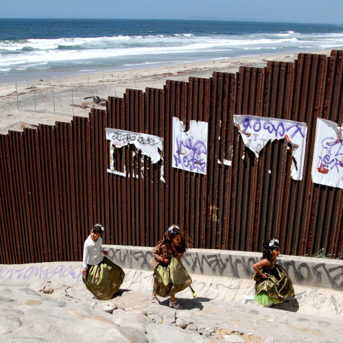 photo of three children in dresses walking along the Tijuana side of the beach border wall next to graffiti and wheat pasted posters