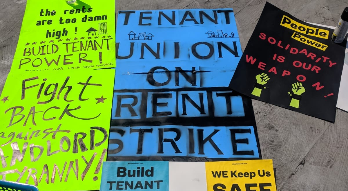 Protest signs reading "the rents are too damn high," "tenant union on rent strike," and "people power - solidarity is our weapon"