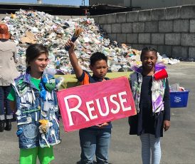 Three schoolchildren in front of a plastic waste dump in Berkeley, California, campaigning against plastic pollution. Child in the center is holding up a "reuse" sign