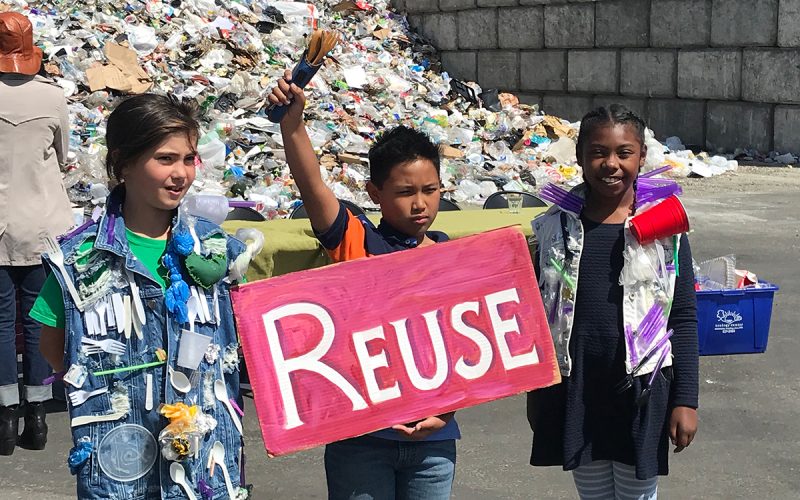 Three schoolchildren in front of a plastic waste dump in Berkeley, California, campaigning against plastic pollution. Child in the center is holding up a "reuse" sign