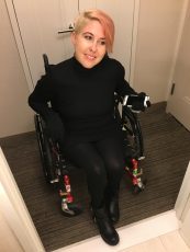 Sydney Pickern> A woman using a wheelchair crosses a threshold with her phone in one gloved hand. She wears black from boots to high necked top. Her hair is short on one side and crosses her forehead on the other. Head tilted to one side, she smiles engagingly. 