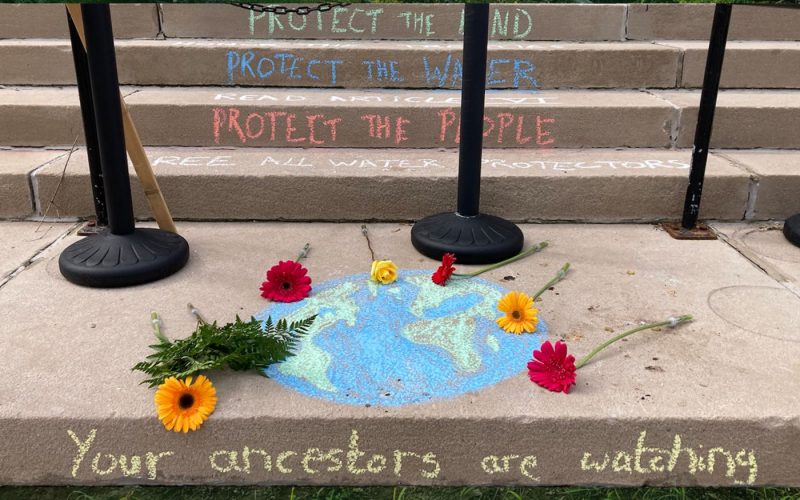 graphic that says in light blue: Water Protector Interview: Jaike Spotted Wolf on Climate, Indigenous Leadership & Solidarity, the light blue painted lady butterfly We Rise logo is on the lower left. The words are overlaid on a photo of tree branches above, and grass and dirt below. In the middle is a photo from the steps of the Aitken County Courthouse, chalk writing on the steps, each statement on a different step, it says: “Protect the Land, Project the Water, Protect the People, Free All Water Protectors”. A chalk drawing of the earth has red and yellow flowers strewn across it. Below that on another steps it says “Your ancestors are watching.”