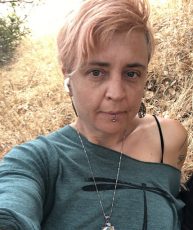 Selfie of shiloh burton with green off the shoulder long sleeve shirt with black dragonfly image, silver necklace & dangly earrings at Olompali State Park in the golden California grass.