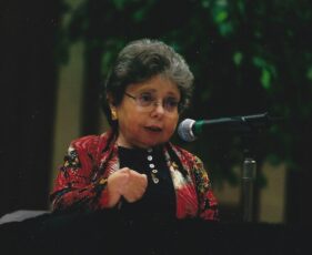 Catherine Campisi. A women speaks into a microphone. She has curly dark hair with a distinguished shock of gray in the front. One of her hands is curled toward her body.