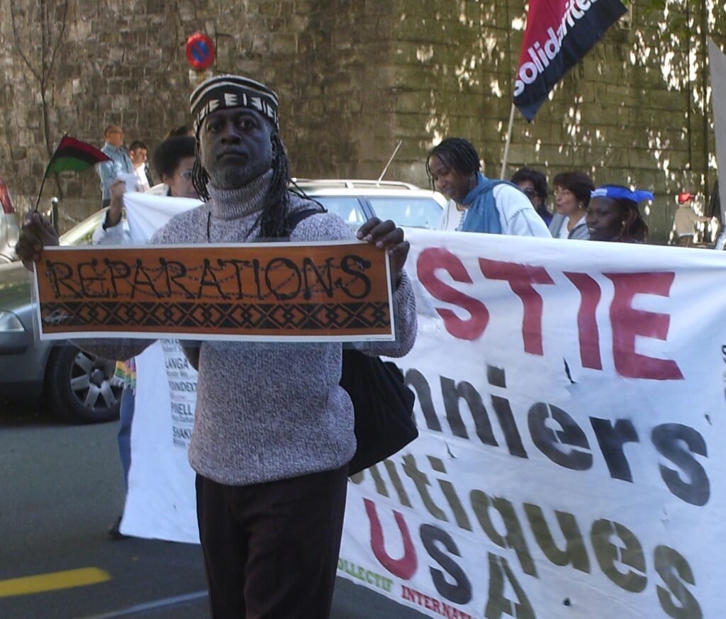 Jahahara Amen-RA Alkebulan-Ma'at marching with the Collectif International Pan Africain at U.N. headquarters in Switzerland, holding a poster of artwork reading "Reparations" by Emory Douglas.