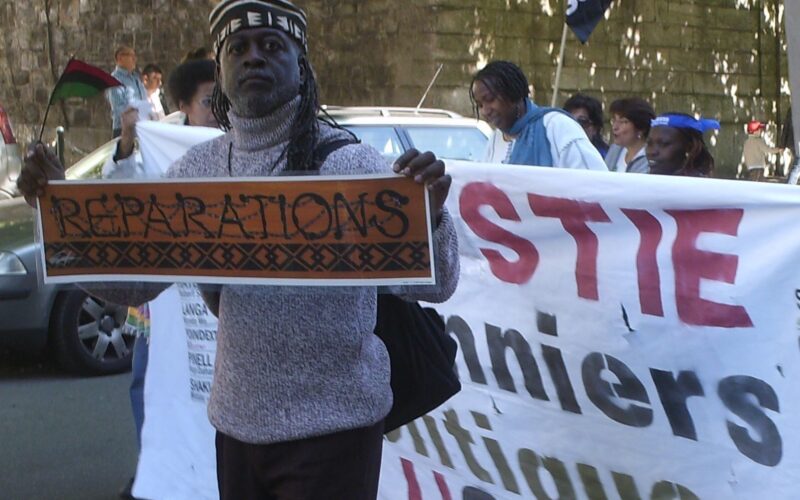 Jahahara Amen-RA Alkebulan-Ma'at marching with the Collectif International Pan Africain at U.N. headquarters in Switzerland, holding a poster of artwork reading "Reparations" by Emory Douglas.
