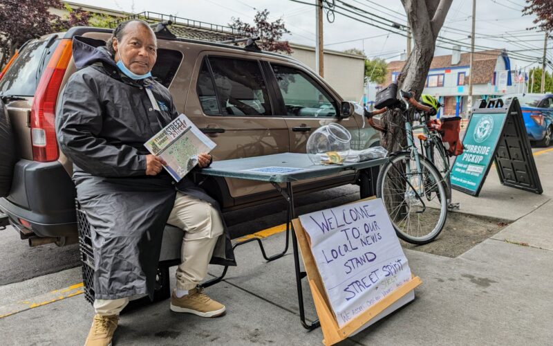 An African American man seated on a sidewalk next to a card table with a stack of newspapers on it next to a clear jar. A hand-lettered sign in front of the table reads "Welcome to our local news stand"