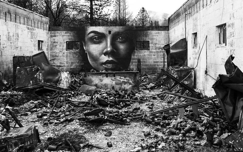 black and white image of a mural showing a woman's face painted on a wall of a burned out building with no roof. Blackened, burned cans and furniture lie in the foreground.