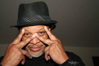 a photo of Ashanti Alston smiling at the camera wearing a pork pie hat and making a mask with his fingers in front of his face